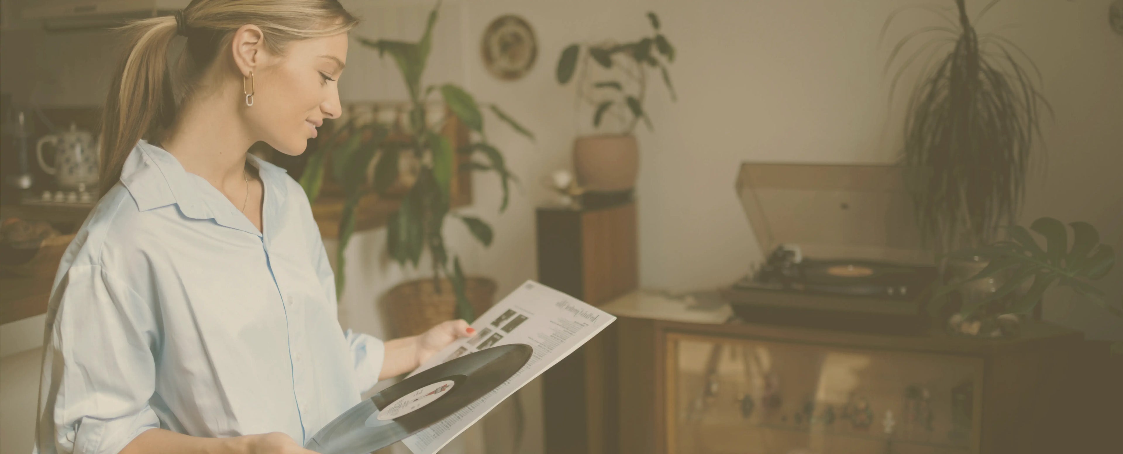 Homepage header image for Peninsula Records and Books, showing a woman in a light blue shirt holding a vinyl record and sleeve in a cosy home setting, with plants and a turntable in the background.