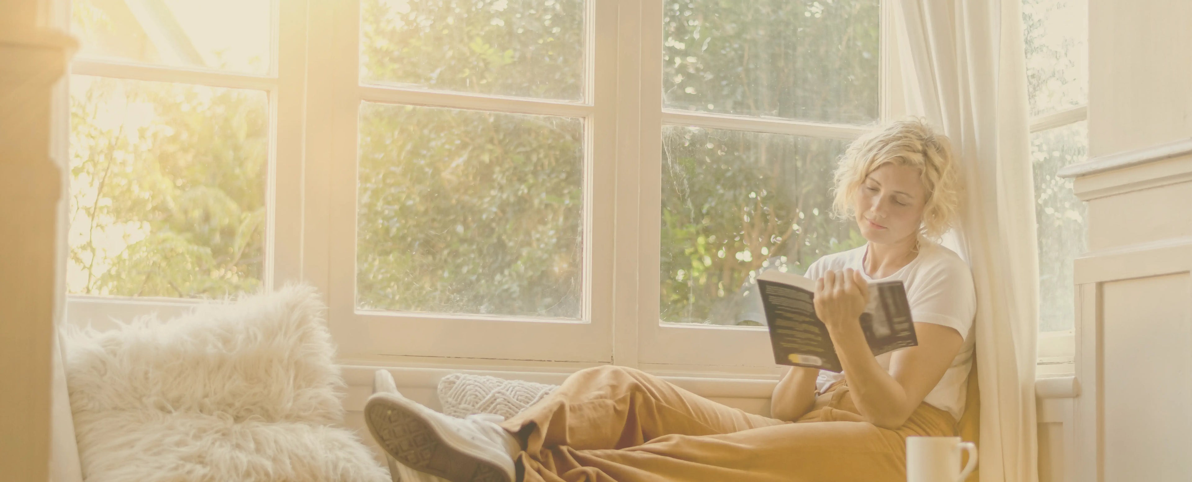 Homepage slideshow image for Peninsula Records and Books, showing a woman sitting by a sunny window nook reading a book, with soft natural light, white curtains, and a cosy cushion beside her