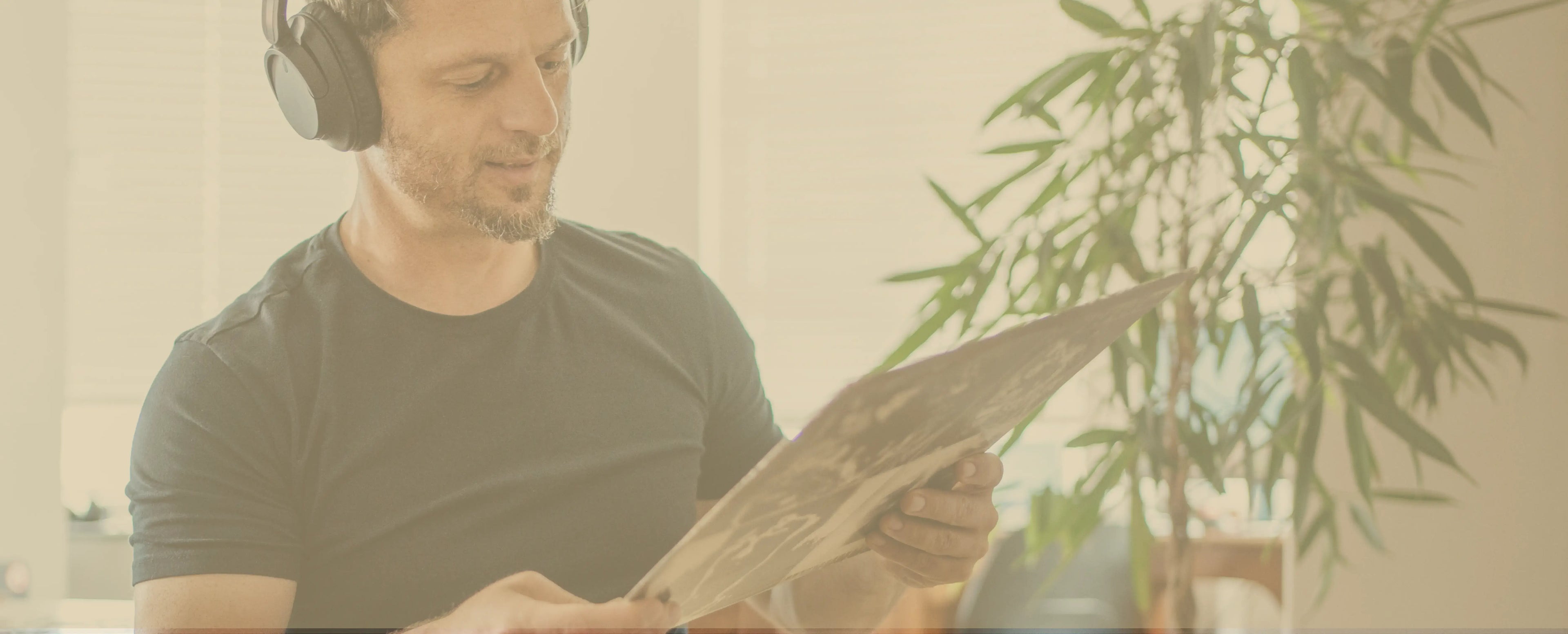 Homepage footer image for Peninsula Records and Books, showing a man in headphones smiling while holding a vinyl record sleeve, with indoor plants and natural light creating a relaxed listening atmosphere.