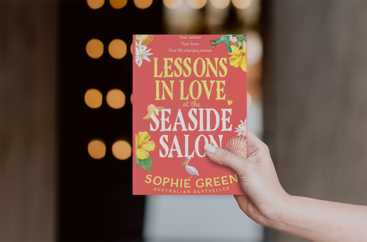 Book titled 'Lessons in Love at the Seaside Salon' held by a hand with a blurred background